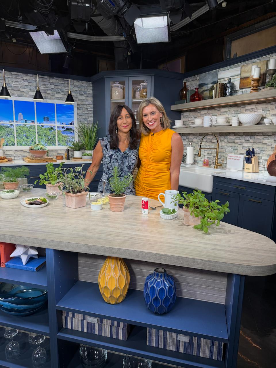 Luisa Szakacs and Eve Russo behind a kitchen island with potted herbs on a WFMZ studio cooking set.